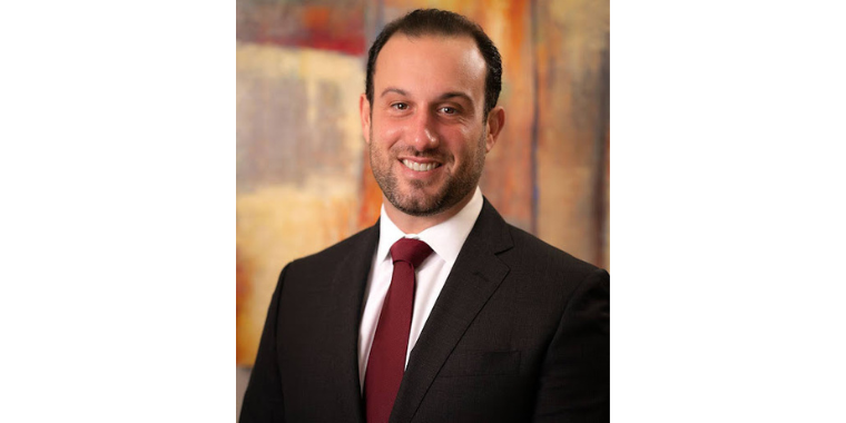 Joseph L. Albano, DO, MBA, smiles in black suit and red tie in front of bright background. 